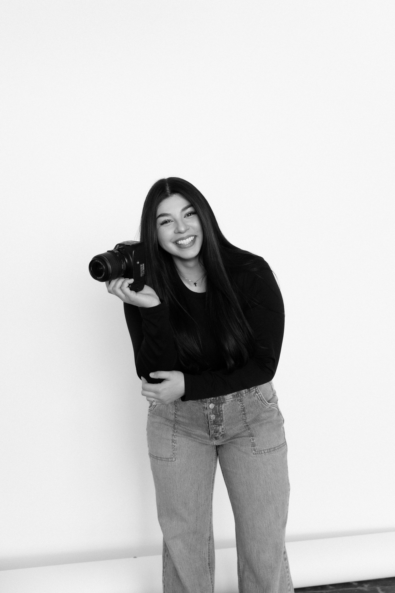 A photographer in black sweater poses playfully with camera against white wall.