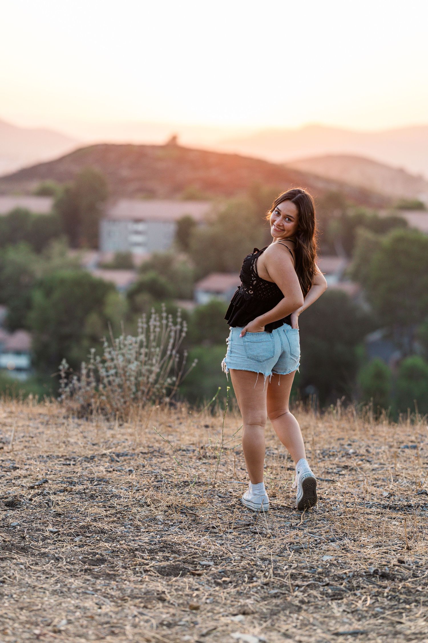 Someone in denim shorts and black top poses on a hilltop during golden hour sunset with mountains in background.