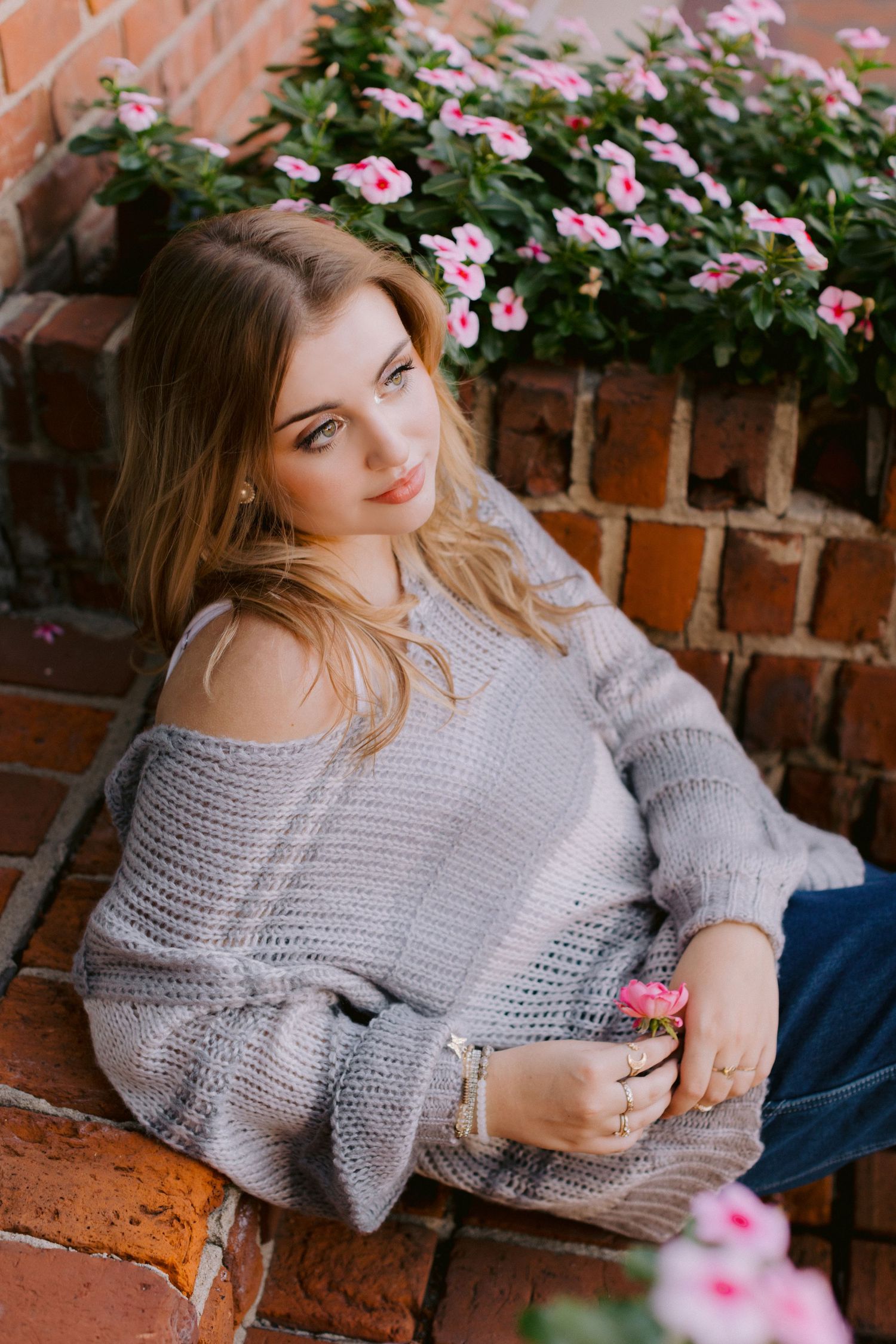 A person in a cozy gray knit sweater sits by a brick wall with pink flowering plants in the background.