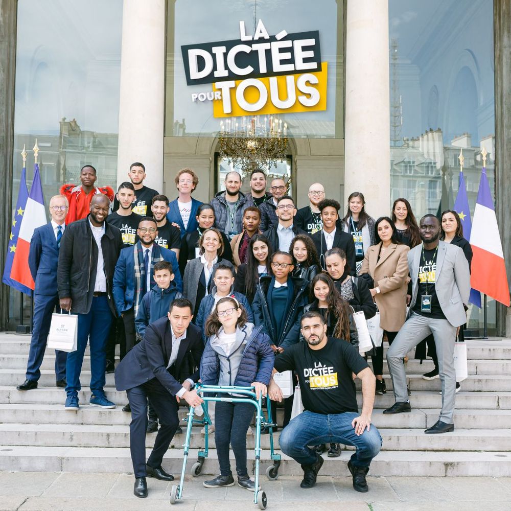 A group of people standing on stairs in front of a building with French flags and a sign that says 'La dictee pour tous'.