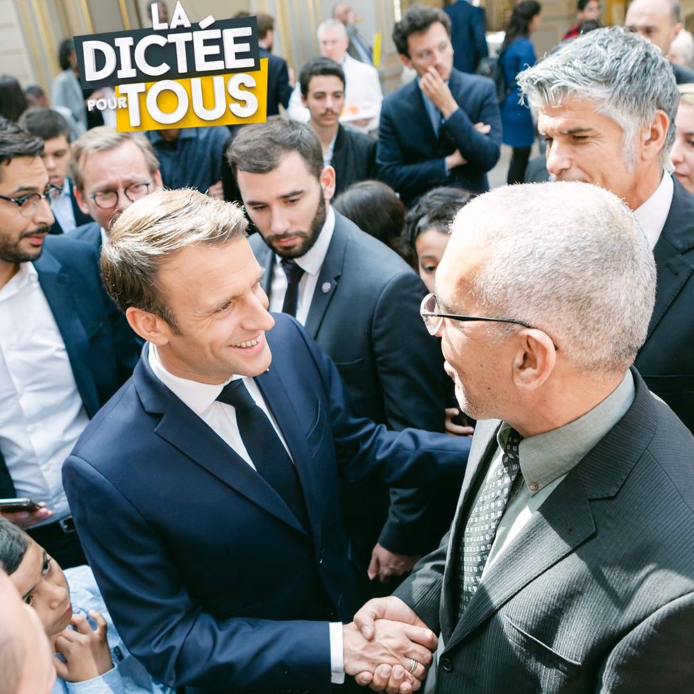 Two men in suits shaking hands during a business gathering with a crowd of onlookers behind them.