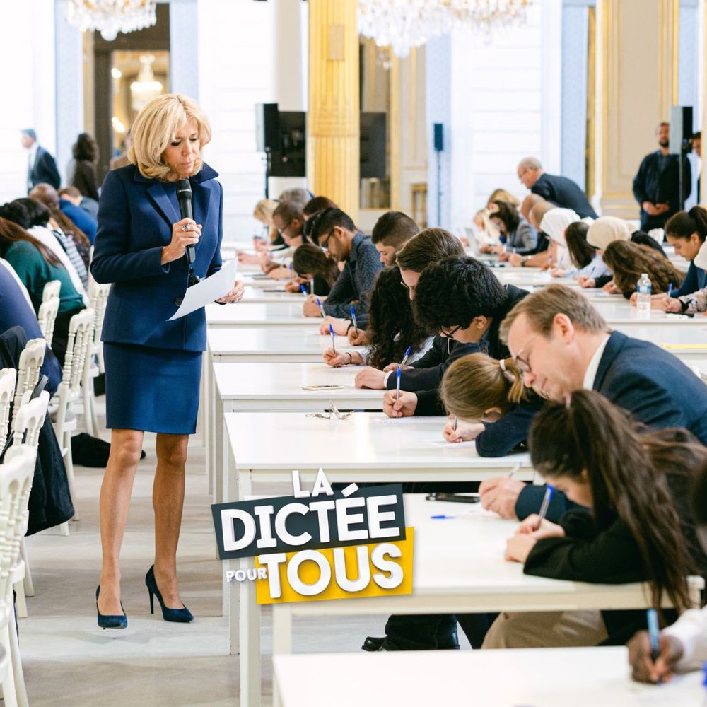 Students write in an exam hall while a teacher in a blue outfit supervises the dictation test.