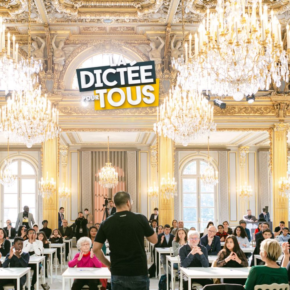 A grand ornate ballroom with crystal chandeliers hosts a formal dictation event with rows of people writing at tables.