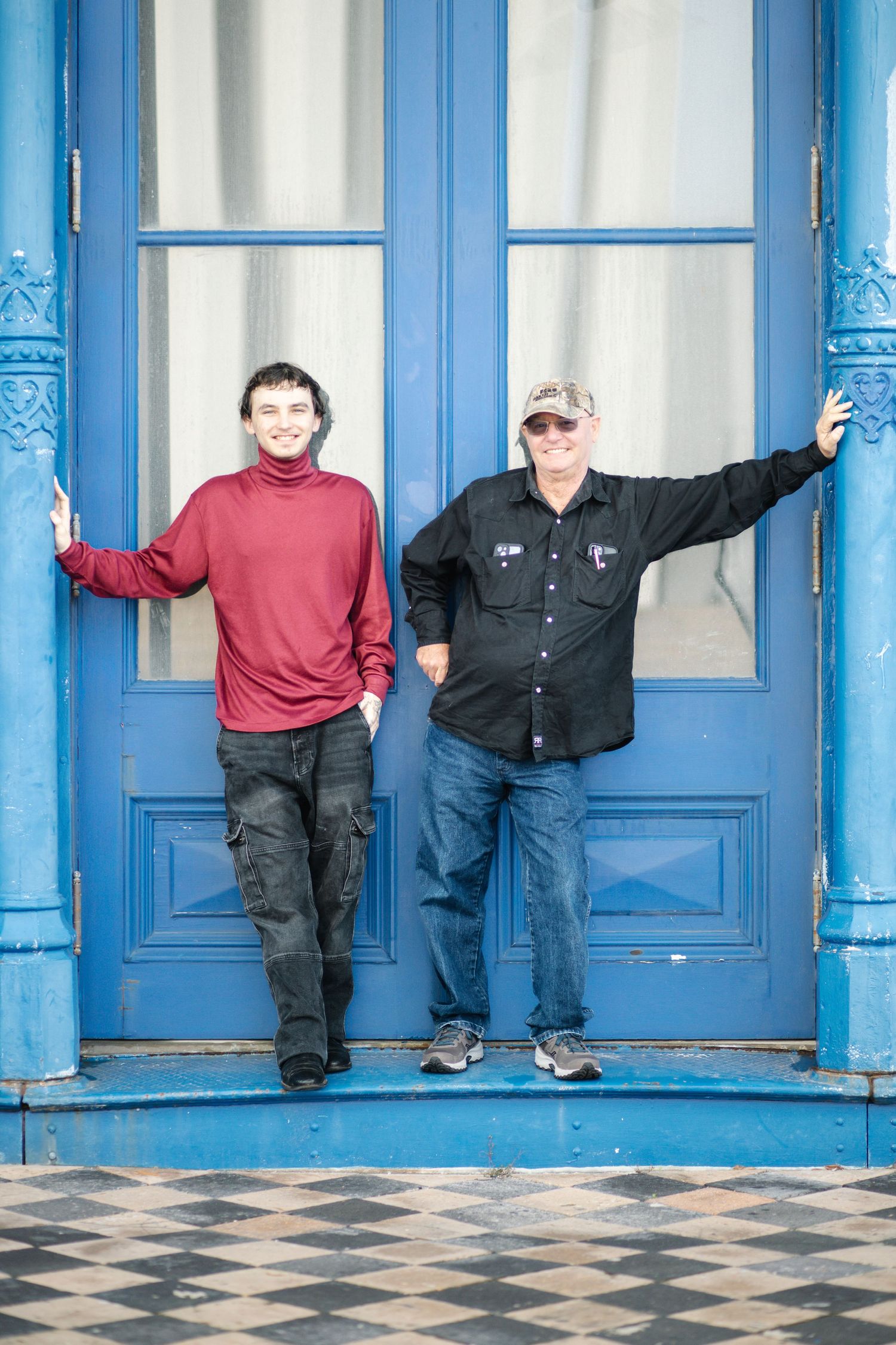 Two people stand in front of bright blue double doors with ornate trim and a checkered floor.