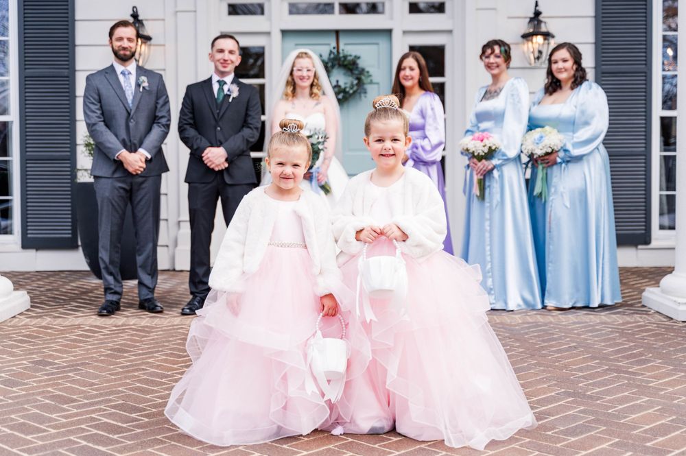 Wedding party poses on brick steps with flower girls in pink tulle dresses at a classic white venue.