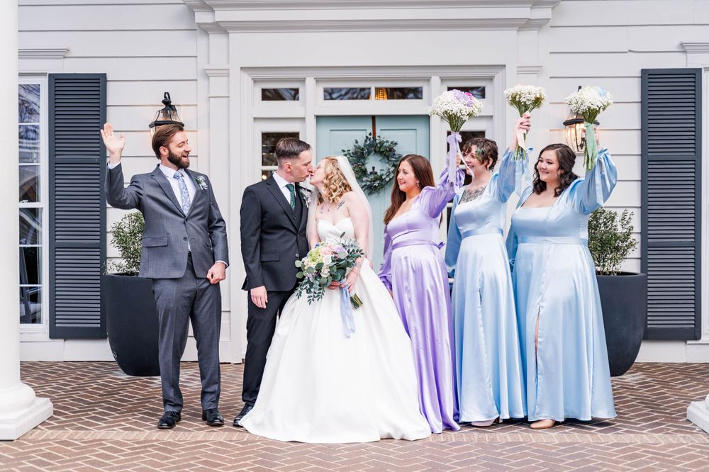 Wedding party in coordinating blue and purple dresses stand together on brick patio.