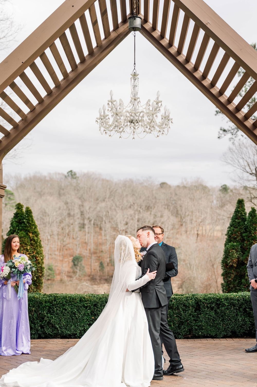 Wedding ceremony under wooden pergola with crystal chandelier and evergreen landscaping. The Highgrove Estate, Fuquay-Varina, NC