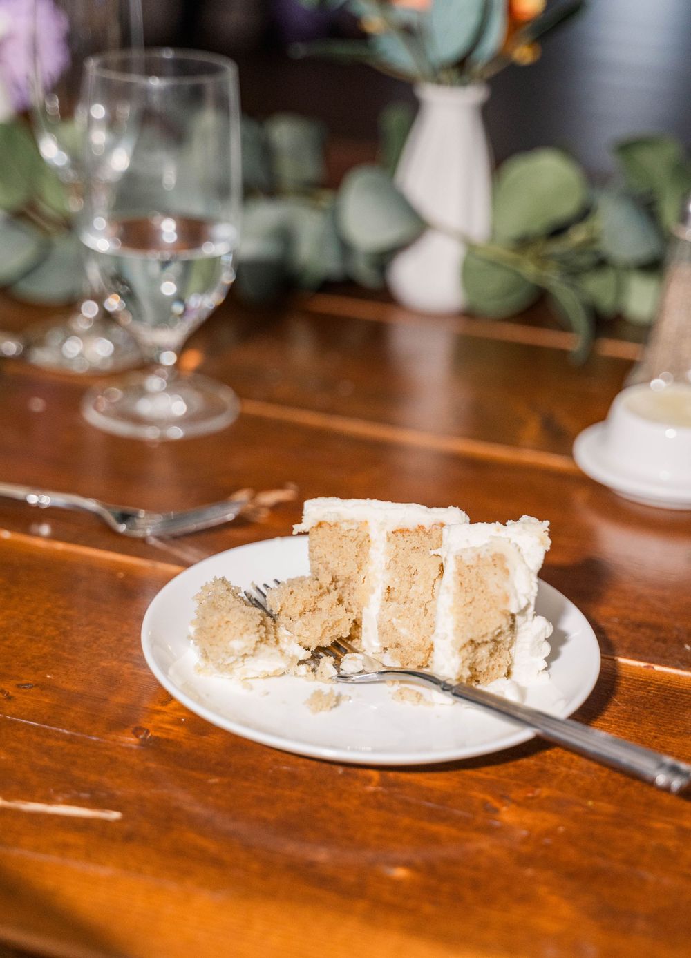A slice of white wedding cake with fork on a wooden table with wine glass and greenery decor.