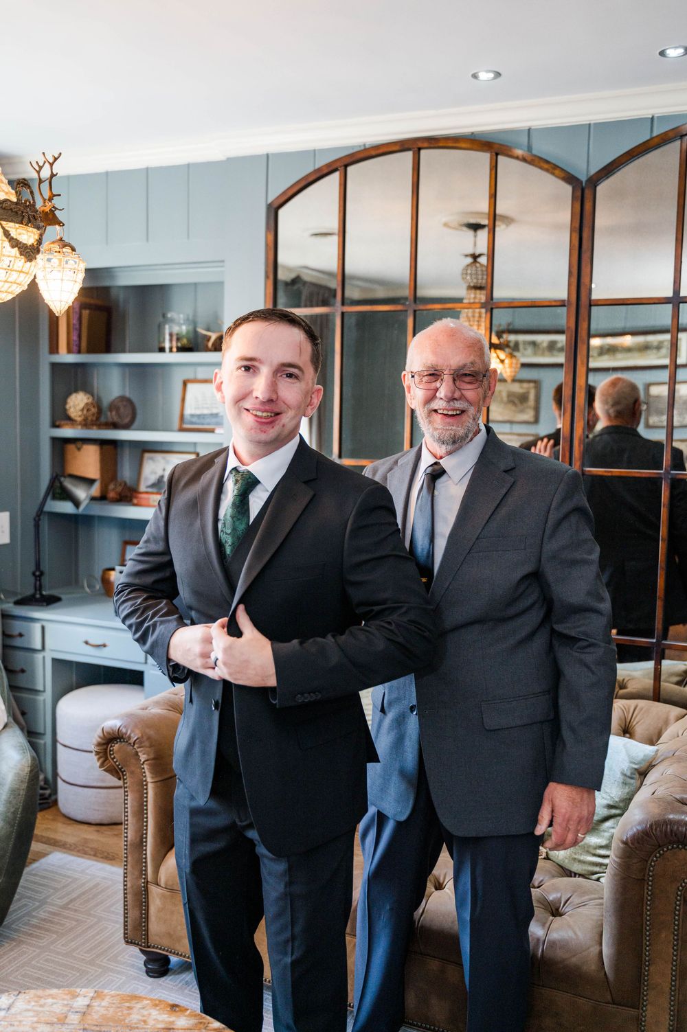 Two men in formal suits standing in an elegantly decorated living room with arched mirrors.