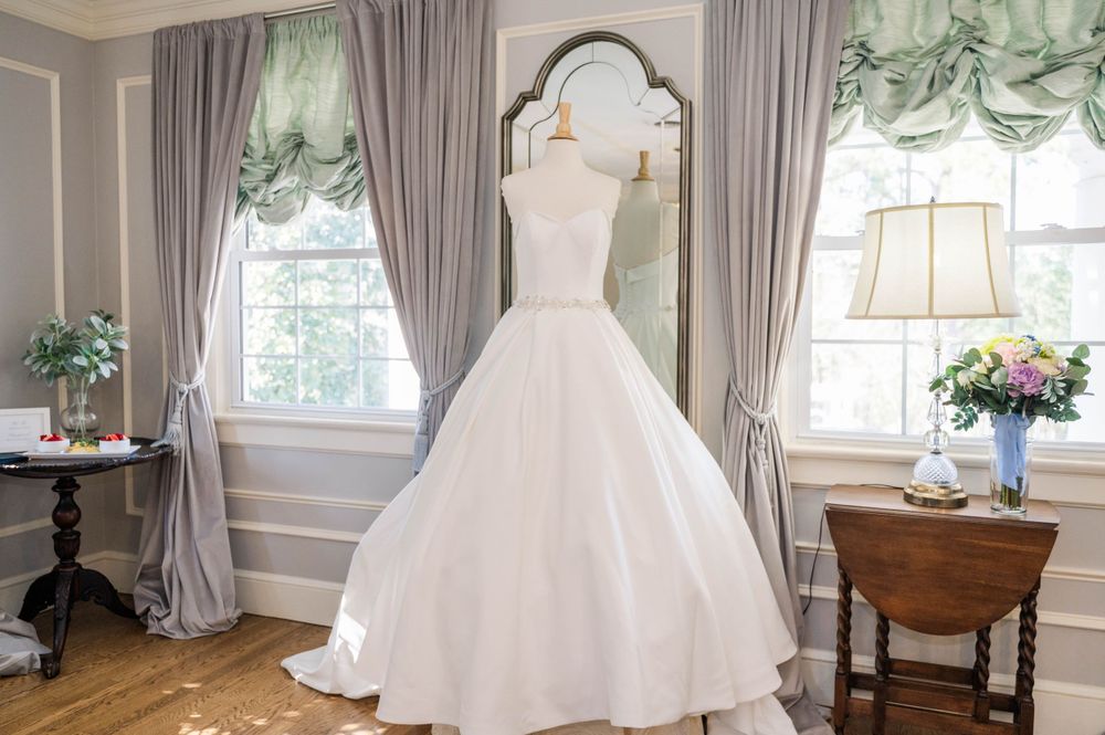 Elegant white wedding dress hanging in a room with gray curtains and vintage decor. The Highgrove Estate, Fuquay-Varina, NC