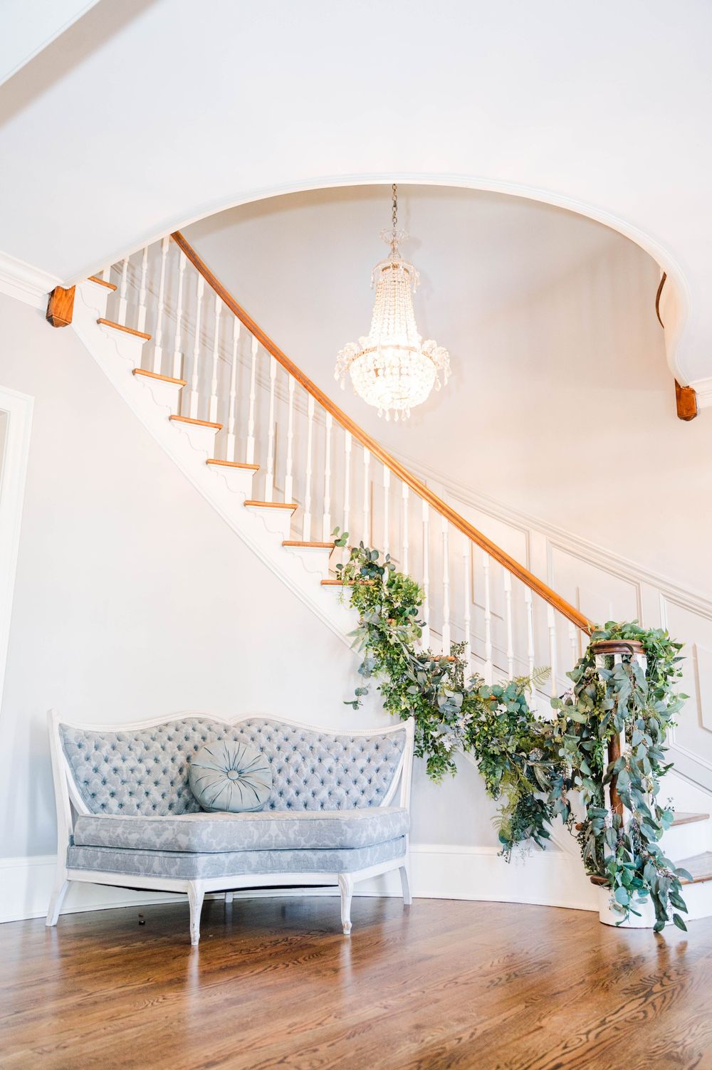 Curved staircase with crystal chandelier and grey settee in bright white interior. The Highgrove Estate, Fuquay-Varina, NC