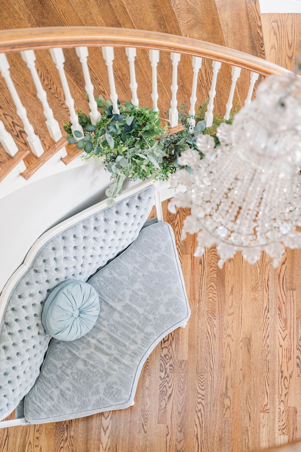 Elegant velvet gray chaise lounge with decorative pillow and greenery under a crystal chandelier on hardwood floors. The Highgrove Estate, F