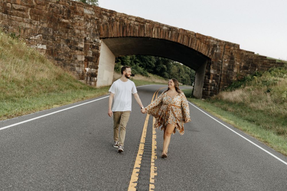A couple walking hand in hand on a road under a stone arch bridge. Floyd, VA