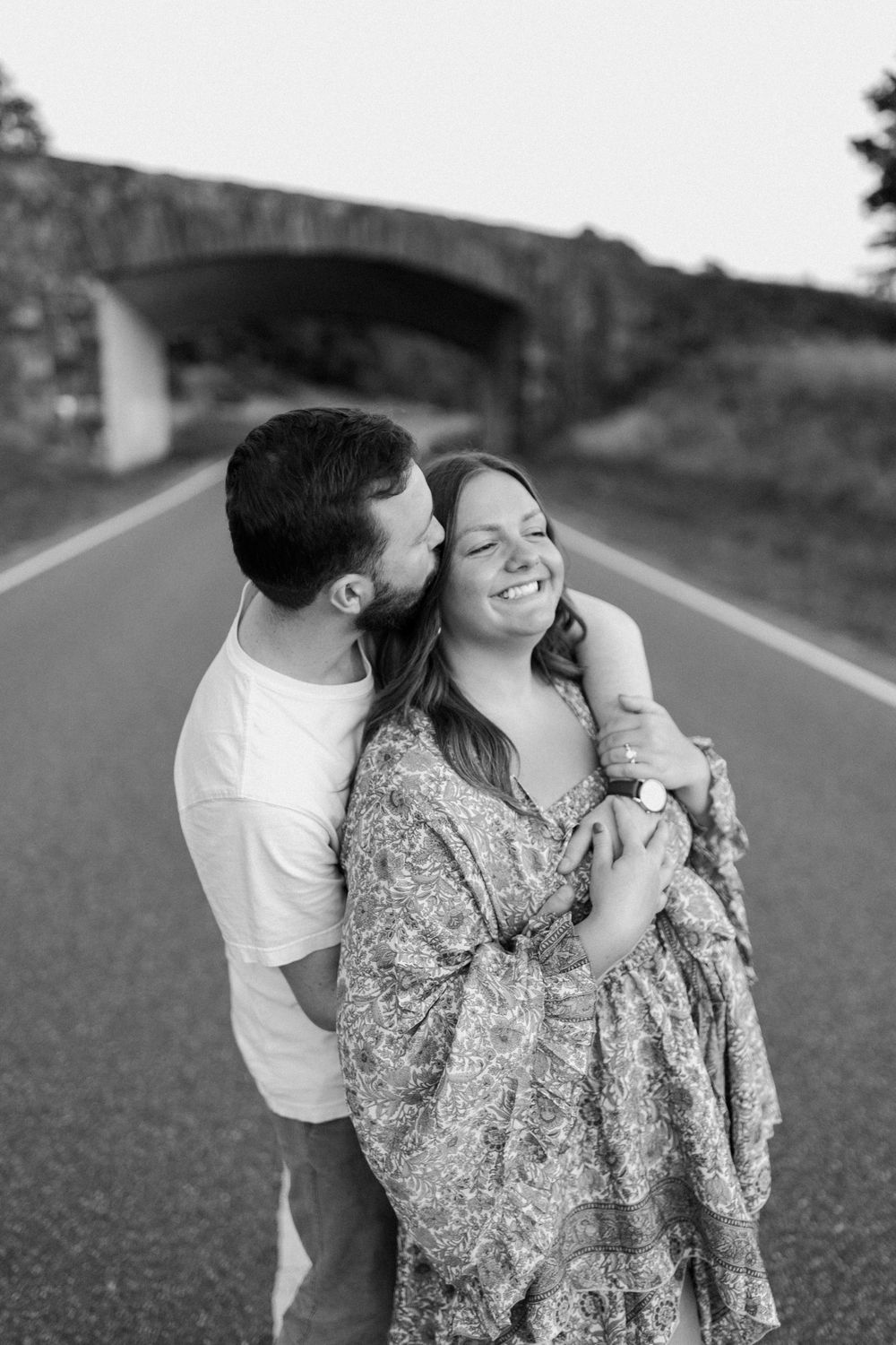 A loving couple embraces on a rural road with stone bridge in background. Floyd, VA