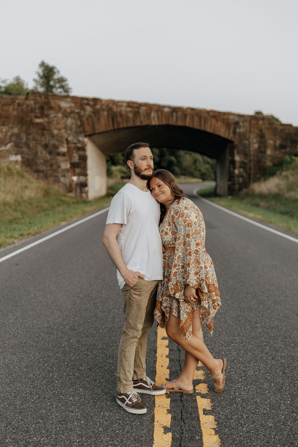 A couple embraces while standing on yellow road lines beneath a stone bridge. Floyd, VA