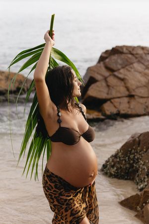 Pregnant woman in brown bikini and leopard print sarong holding palm leaf on rocky beach.
