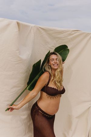 Smiling blonde woman in brown crochet bikini top and mesh skirt holding tropical leaf against beige fabric backdrop.