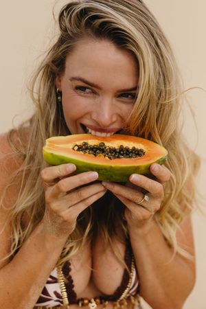 Smiling blonde woman biting into a fresh halved papaya, showcasing vibrant orange flesh and black seeds.