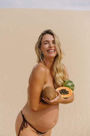 Smiling blonde woman in bikini bottoms holding tropical fruits including papaya, coconut, and watermelon on a beach.