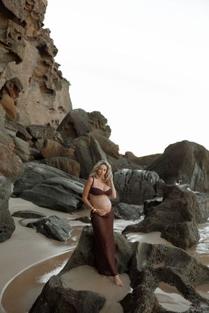 Pregnant woman in burgundy dress posing on rocky beach with large stone formations and ocean waves.