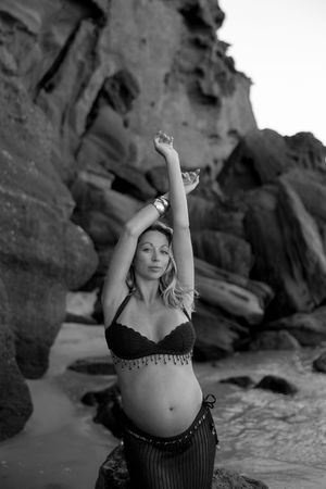 Black and white photo of a pregnant woman in bikini top posing gracefully on rocky beach with arms raised.
