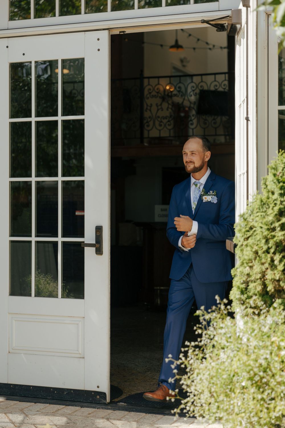 Groom in a fitted blue suit stands in a doorway surrounded by greenery.