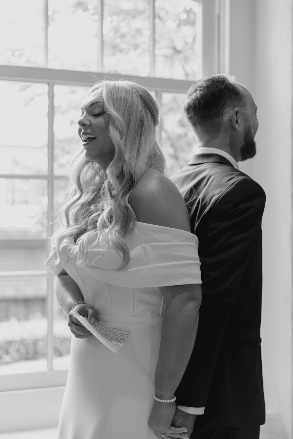 Black and white portrait of a bride in an off-shoulder gown laughing while standing back-to-back with groom.