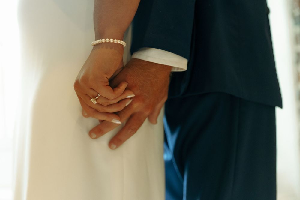 Close-up of hands with wedding rings holding each other against formal attire.