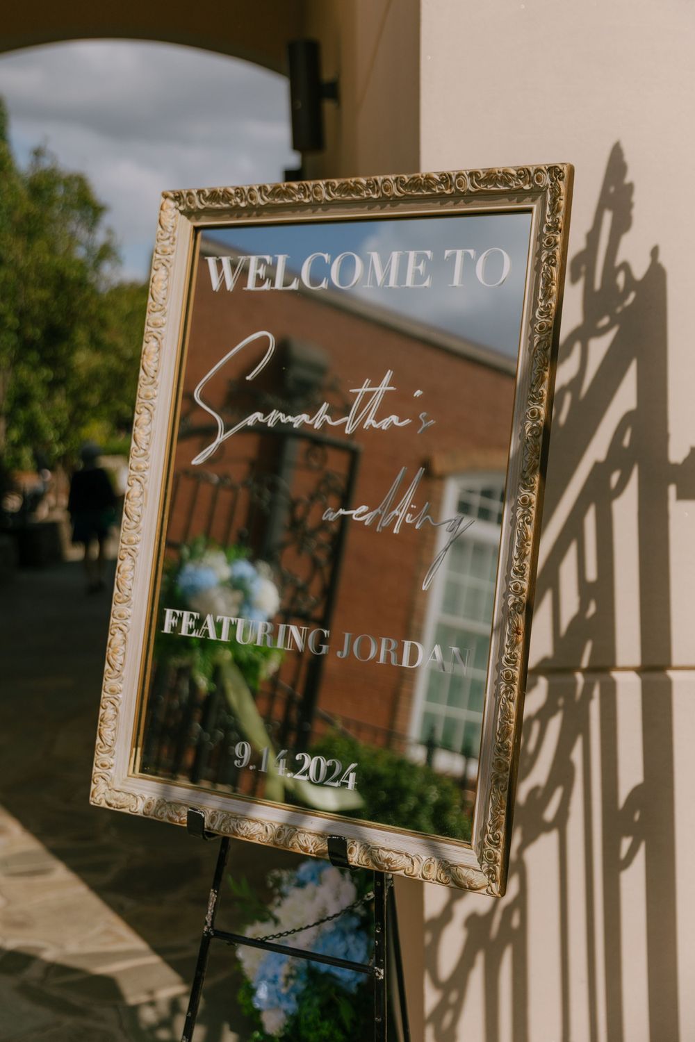 Elegant wedding welcome sign in a gold frame displayed on an easel outdoors.