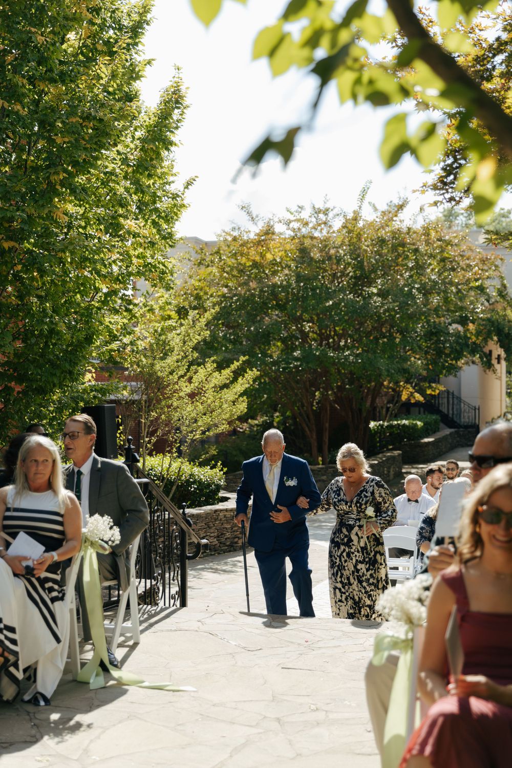 Wedding party members walking down an outdoor pathway lined with trees and white chairs.