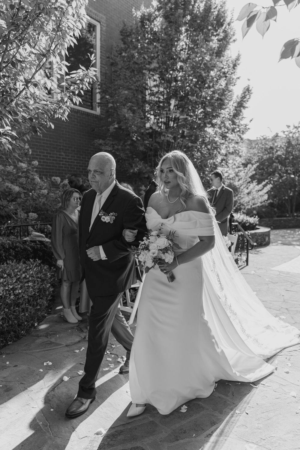 Black and white photo of a bride walking down the path with a bouquet.