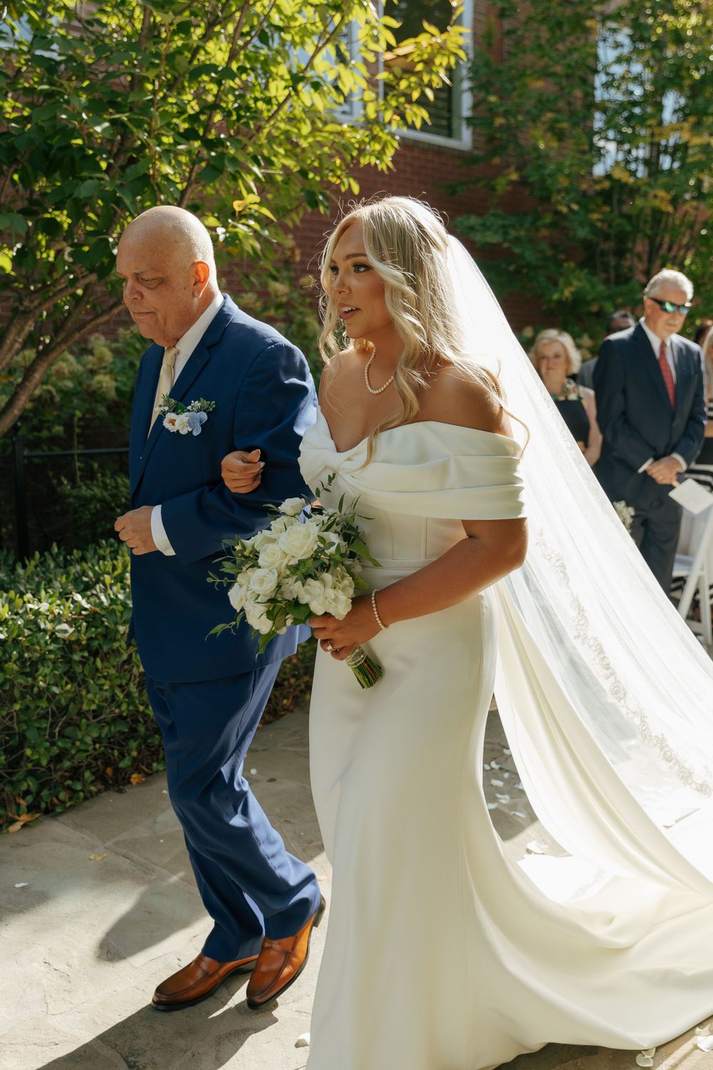 Elegant outdoor wedding procession with bride in flowing white off-shoulder gown escorted down stone pathway.