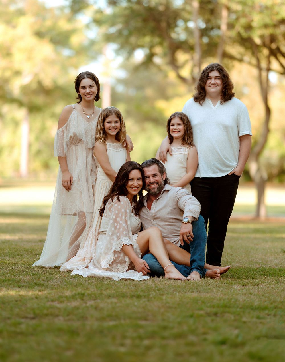 A family wearing coordinated cream and white outfits poses together on grass in a warm sunlit outdoor setting.