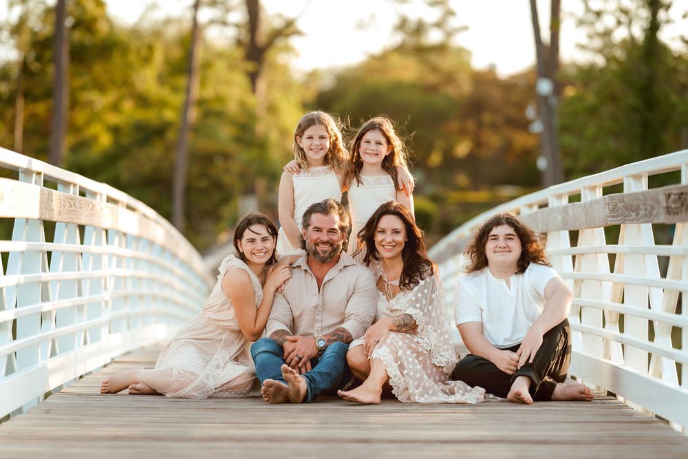 A family poses on a wooden bridge, dressed in neutral tones, with trees and golden sunlight in the background.