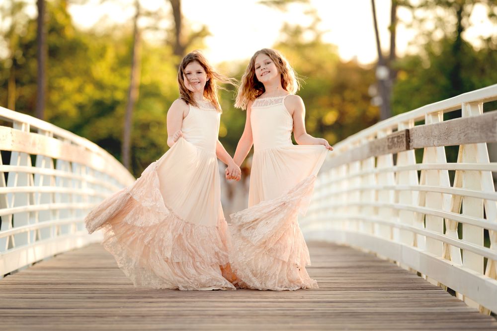 Two girls in flowing blush pink dresses hold hands and laugh while standing on a white wooden bridge at sunset.