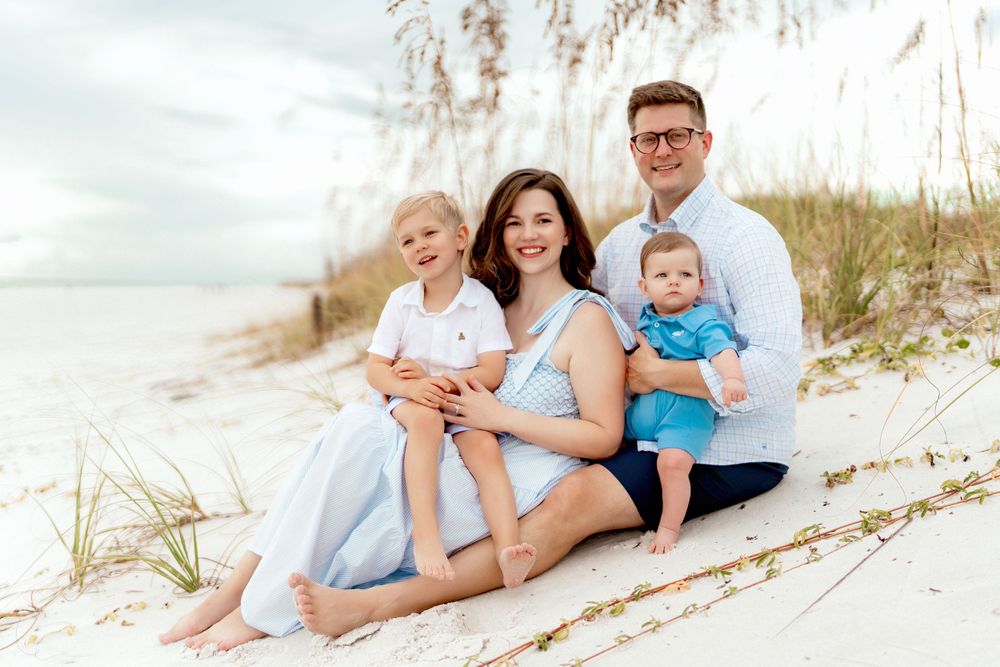 A family sits together on a white sandy beach with sand dunes and sea oats in the background during golden hour.