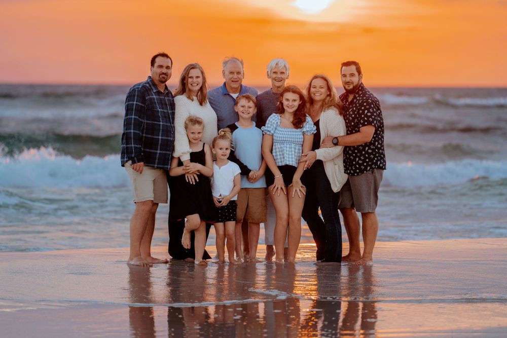 A large family poses together on the beach at sunset, standing in the wet sand with ocean waves behind them and a golden sky above.