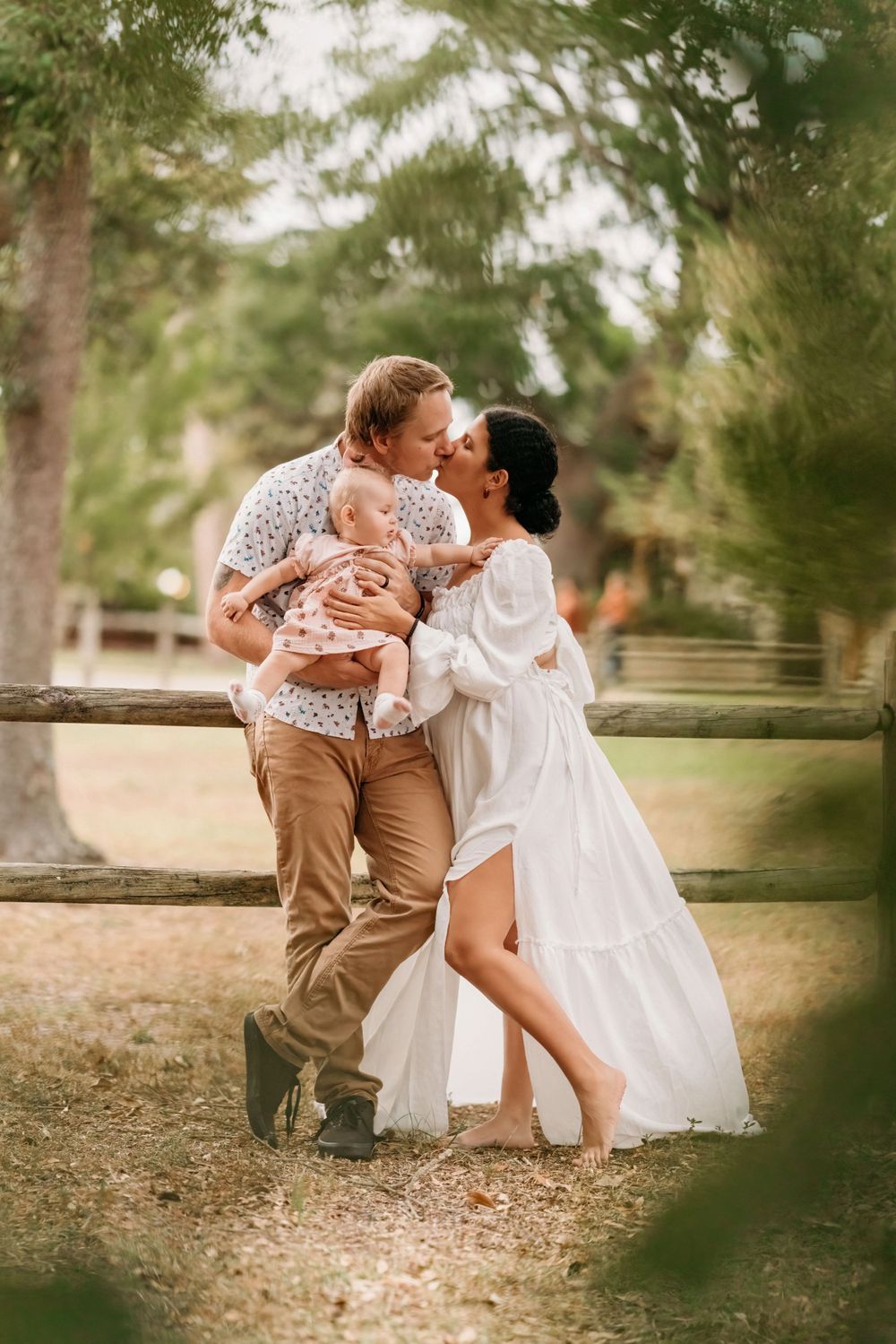 A couple shares a kiss while holding their baby in a warm, outdoor setting with trees and a rustic fence in the background.