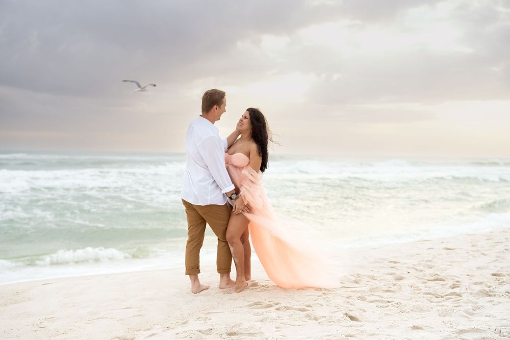 A couple stands barefoot on the beach, embracing as the wind lifts the mother-to-be’s flowing gown, with waves and seabirds in the backgroun
