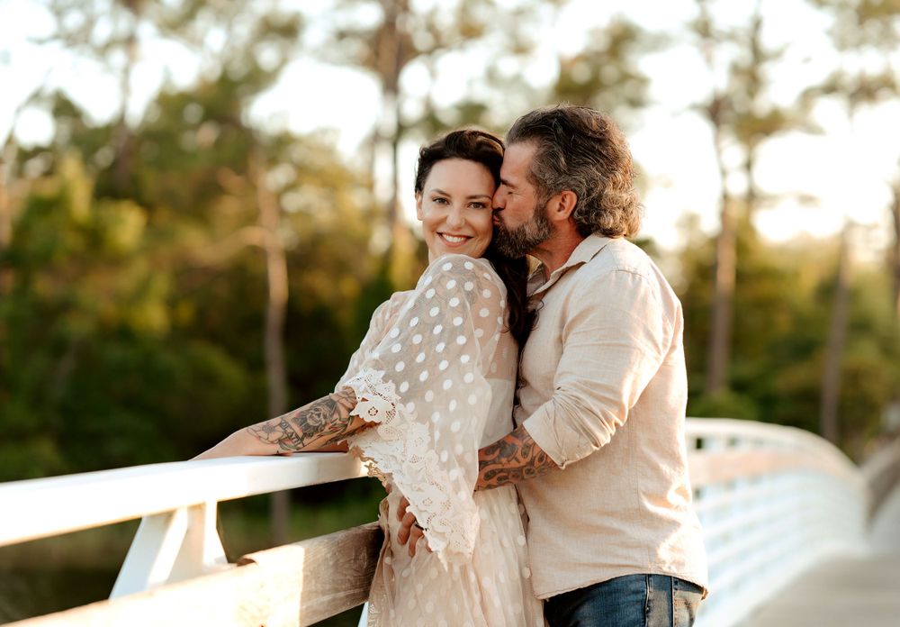 A couple embraces on a wooden deck at sunset, with pine trees creating a warm golden backdrop in the background.