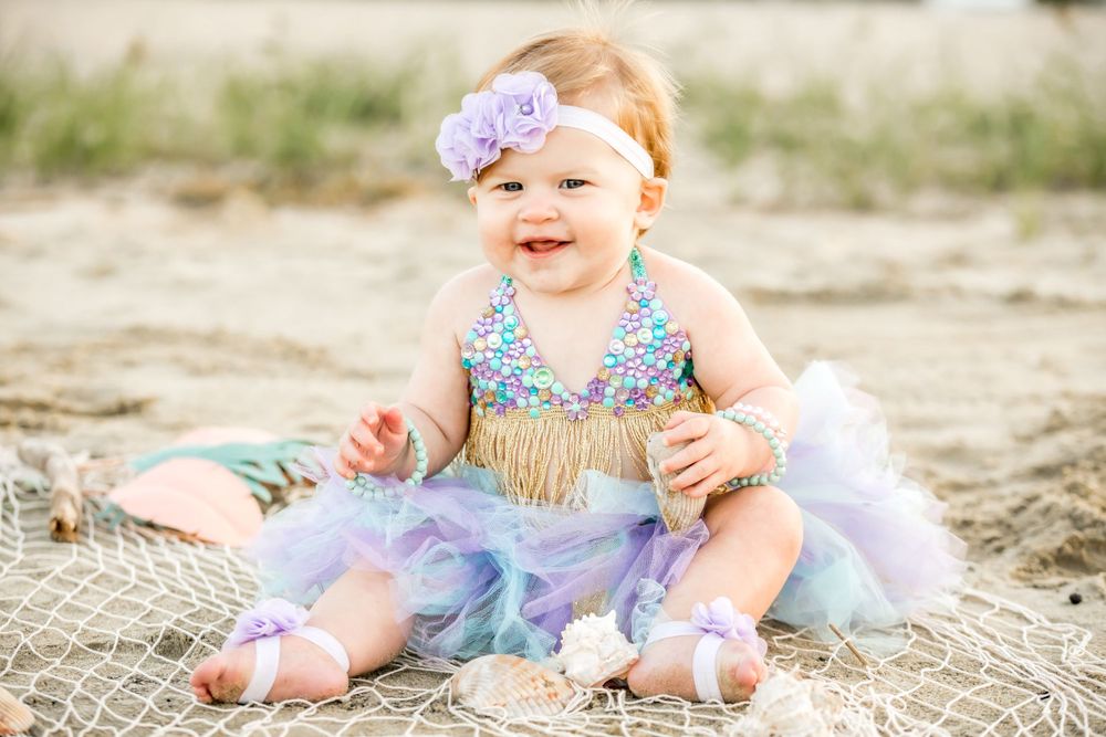 Baby in purple and turquoise tutu dress explores sandy beach setting.