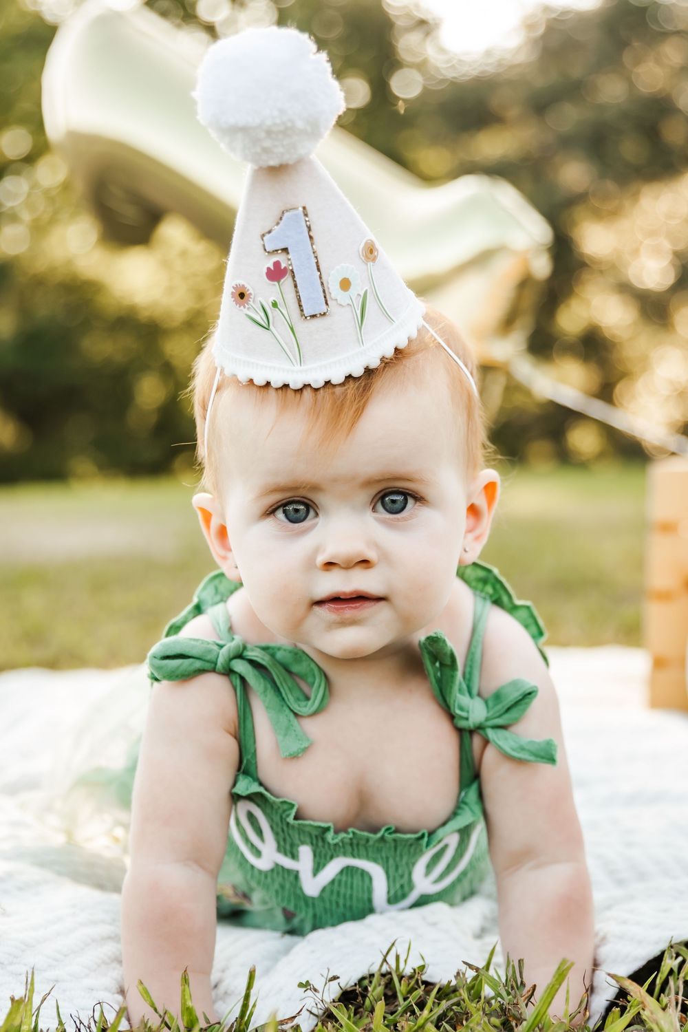Baby wearing green outfit and party hat sitting on white blanket outdoors.