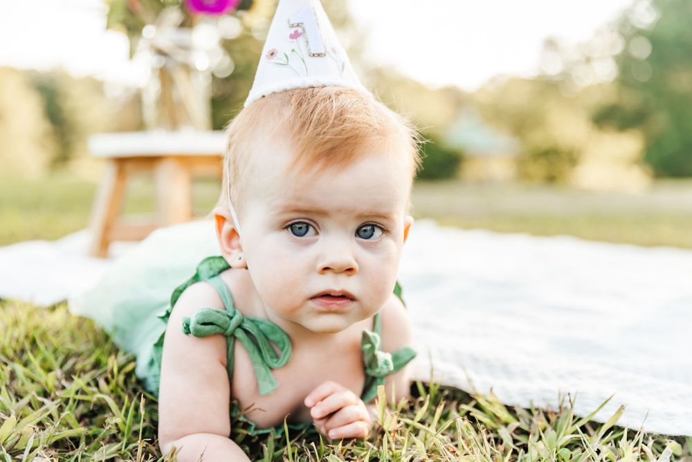 Baby in green outfit and party hat lying on white blanket during outdoor photoshoot.