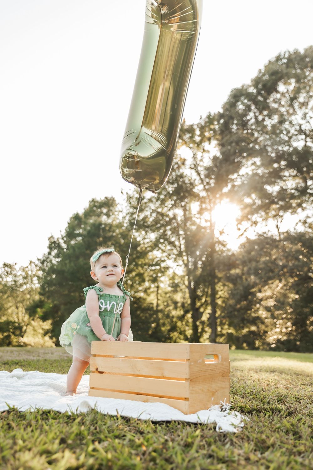 Baby in green romper standing by wooden crate with silver balloon in sunny outdoor setting.