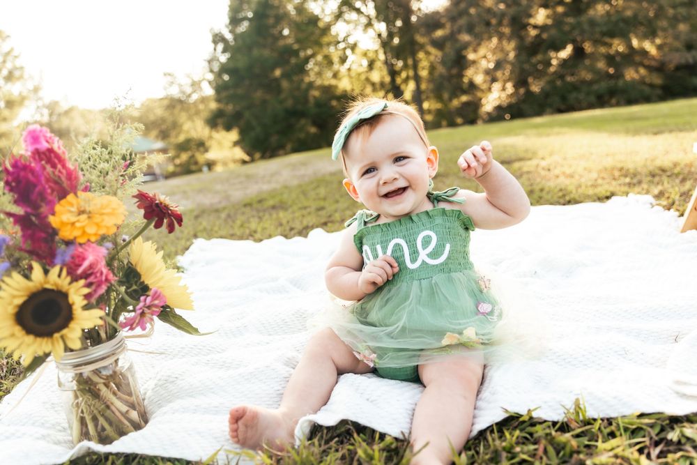 Baby in mint green outfit sitting on white blanket with sunflowers nearby.