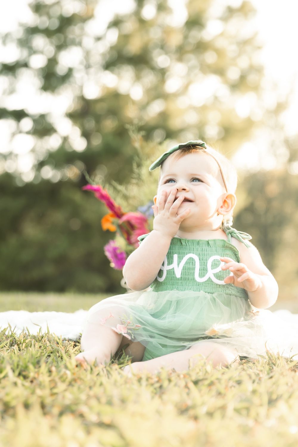 Infant lying on grass laughs while wearing mint green outfit in sunlit outdoor setting.