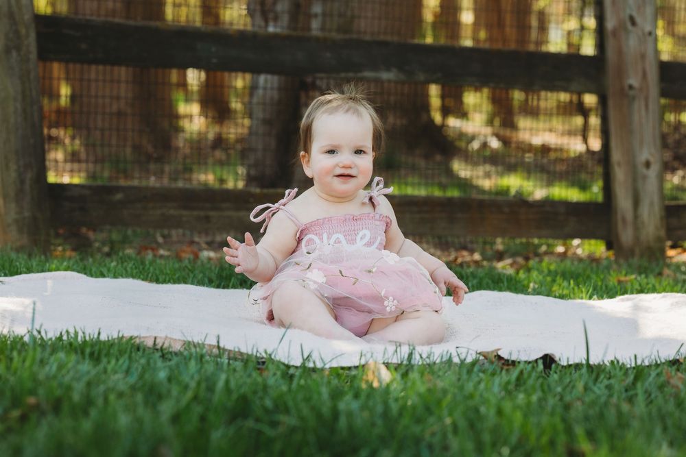Baby girl in pink dress sitting on white blanket in green grass yard smiling outdoors.