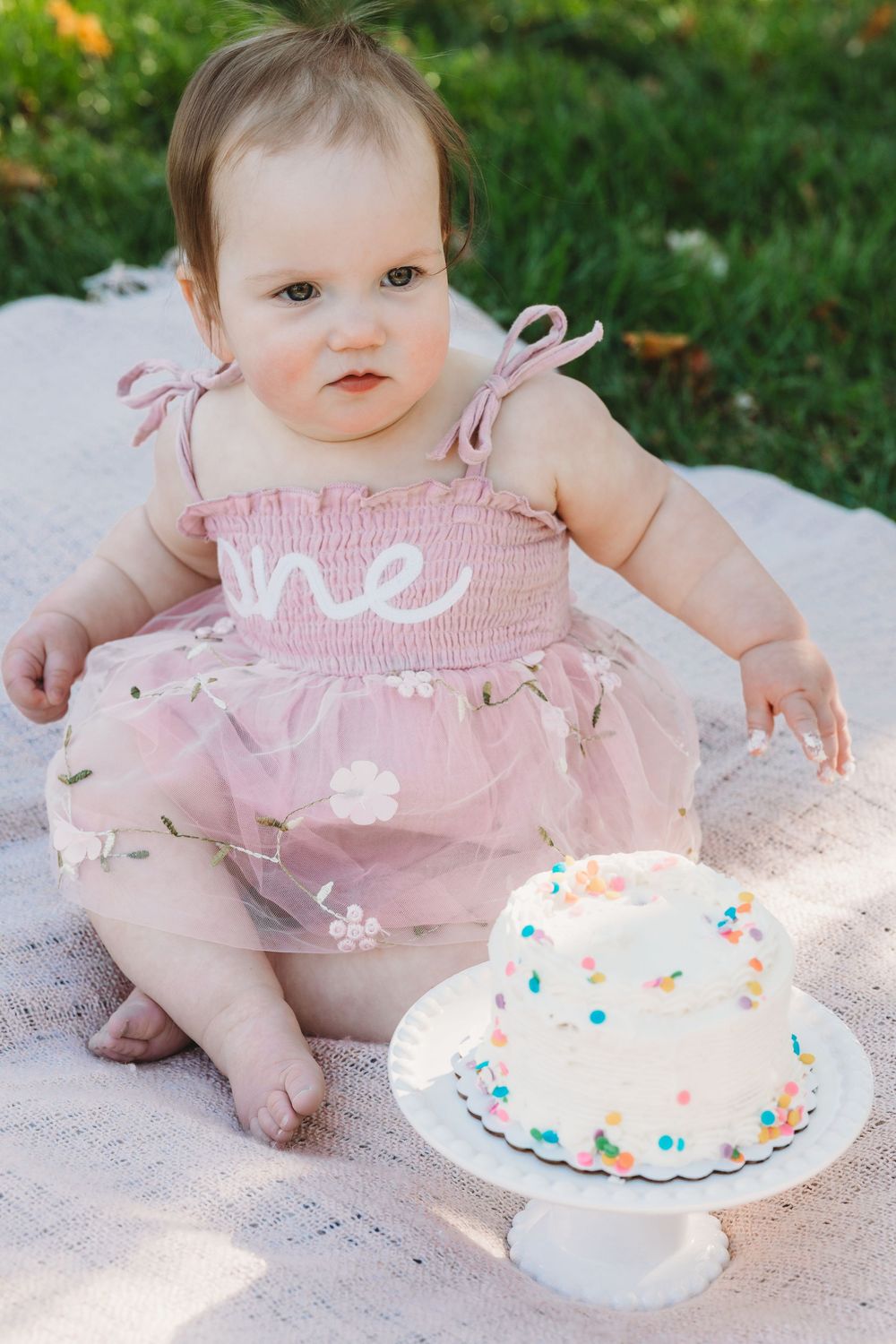 Baby girl in pink tutu dress sits on blanket beside white confetti smash cake for first birthday photo.