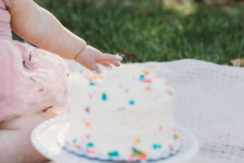 Baby girl in pink tutu touching white sprinkle birthday smash cake outdoors on white blanket.