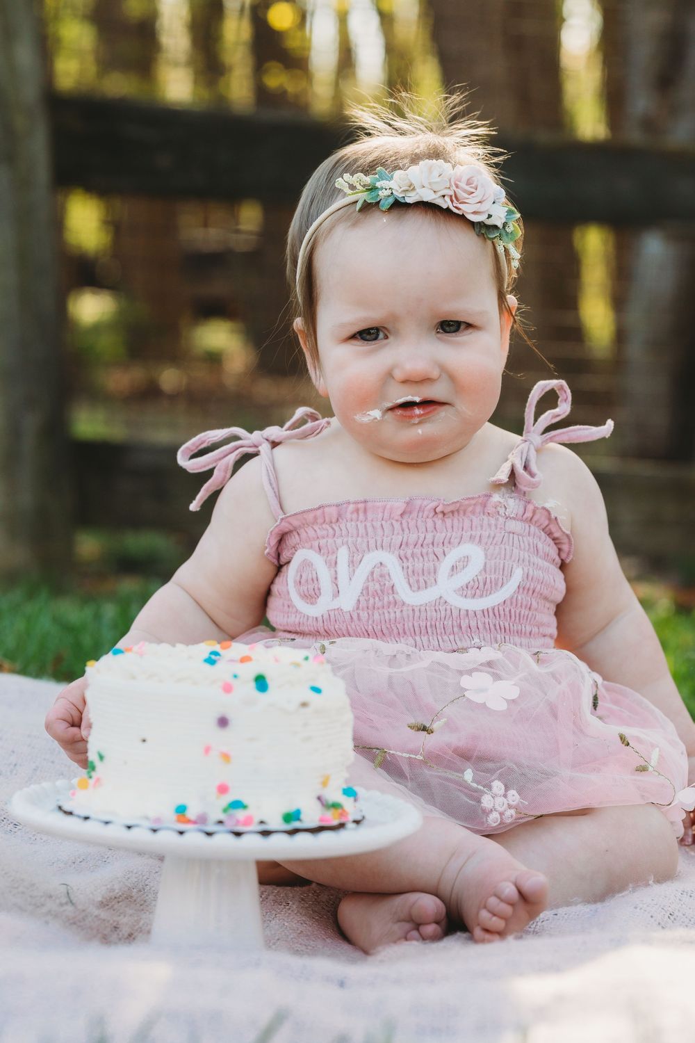 Baby girl in pink dress with floral headband celebrates first birthday with white confetti smash cake outdoors.